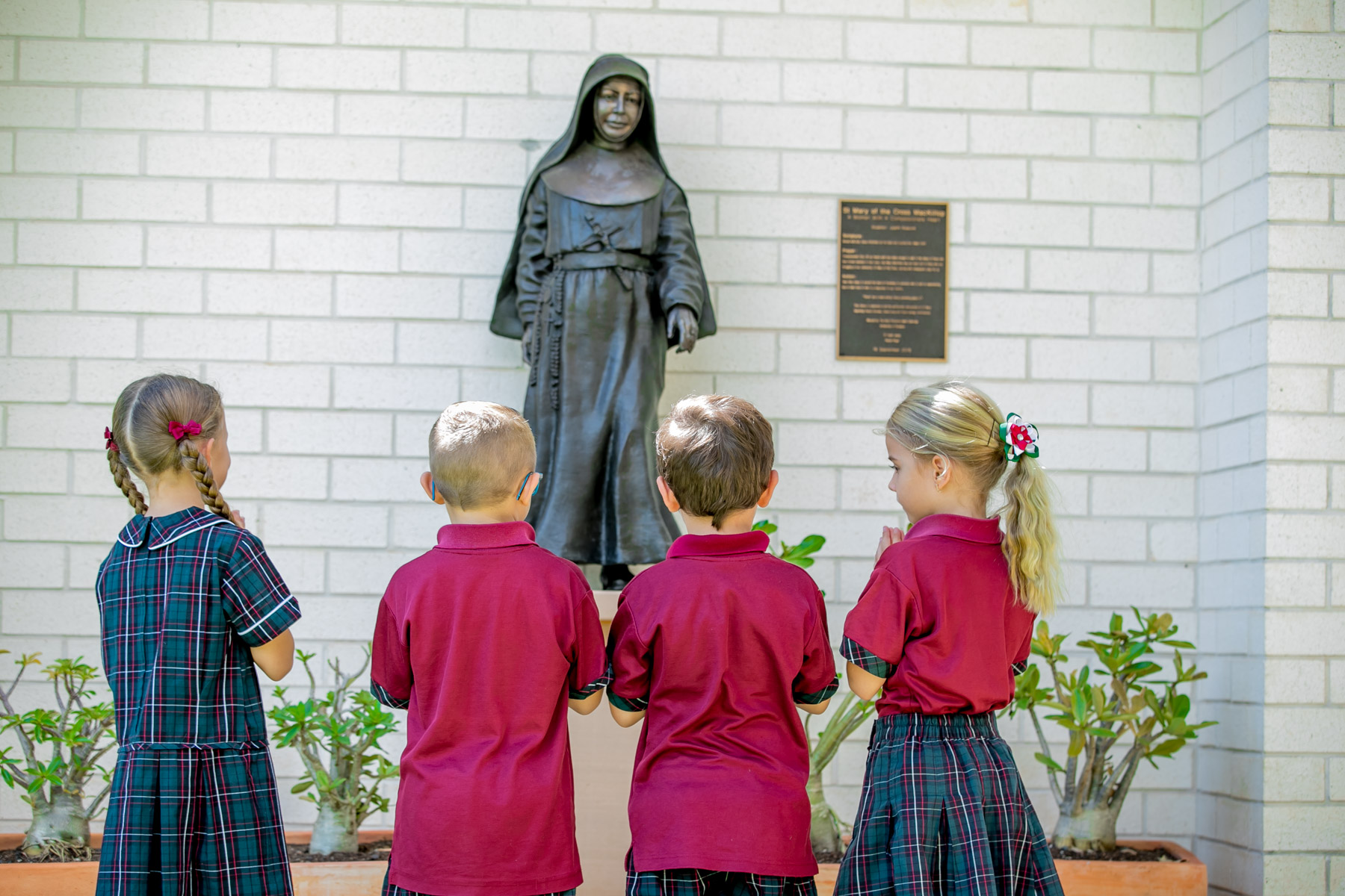 Four primary school students stand facing a statue of a religious sister mounted on a brick wall, with a commemorative plaque beside the statue.
