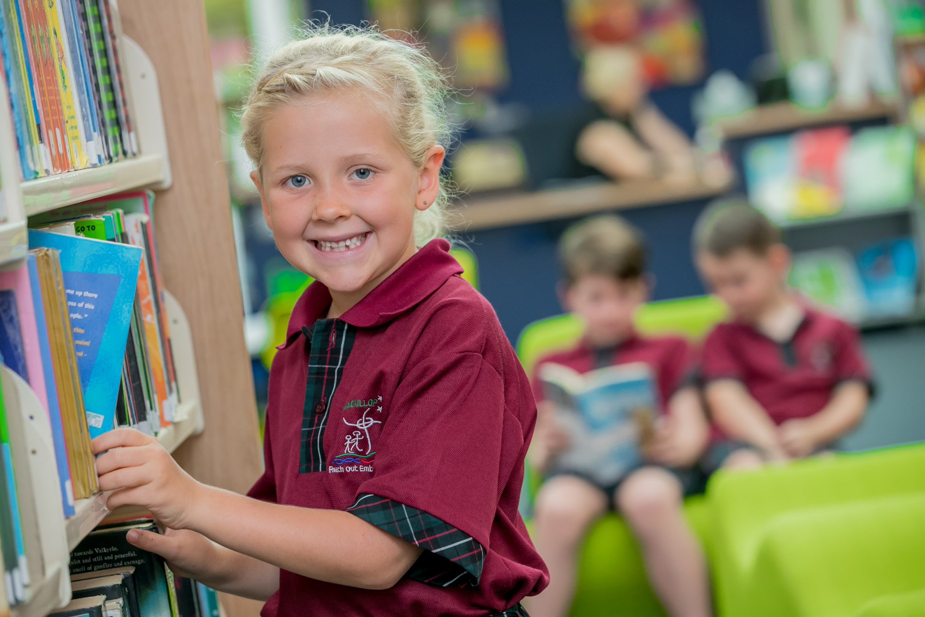 A child taking a book from the shelf. In the background two students are sitting and reading.