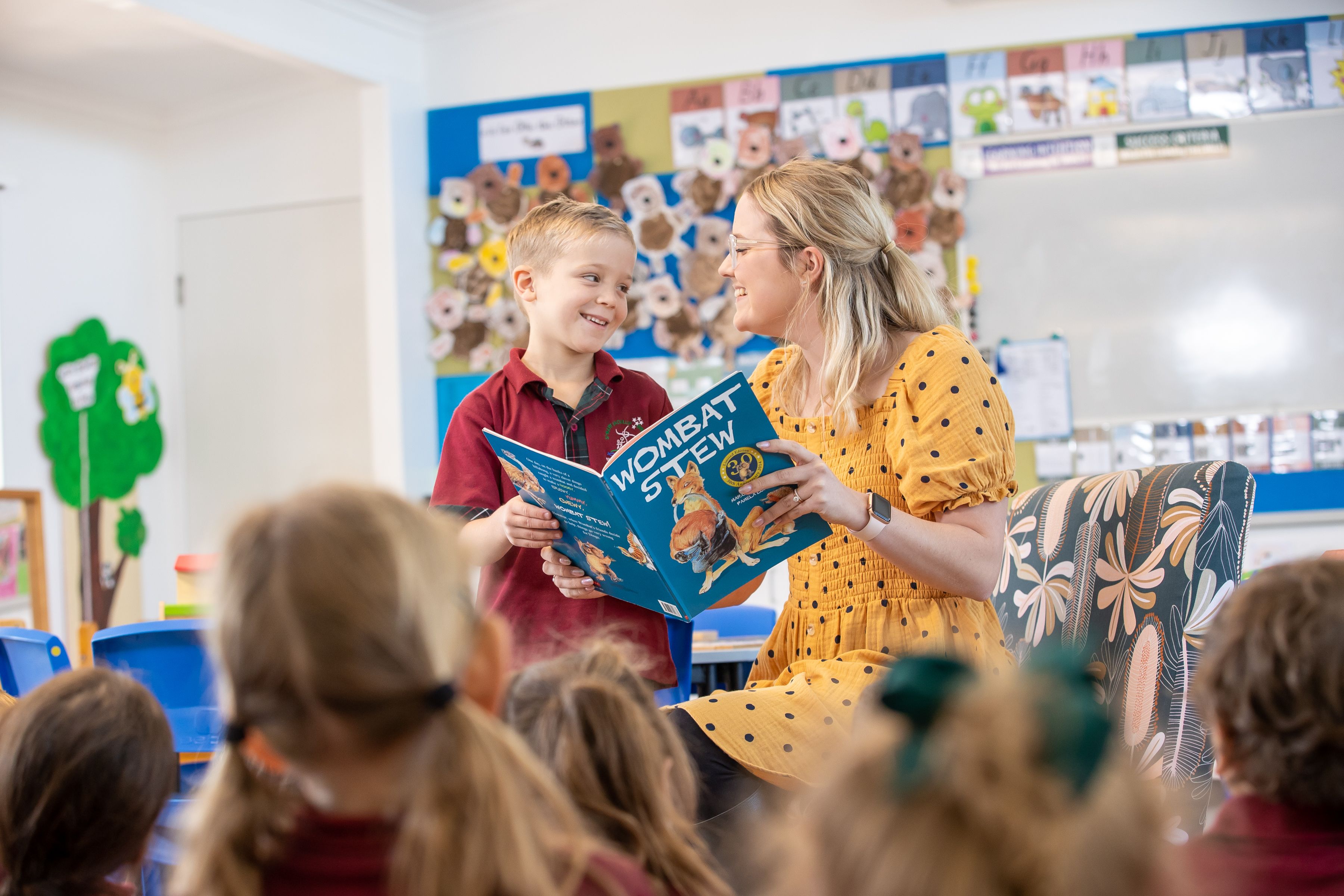 A teacher and a student are holding a book. They are standing in front of the classroom.