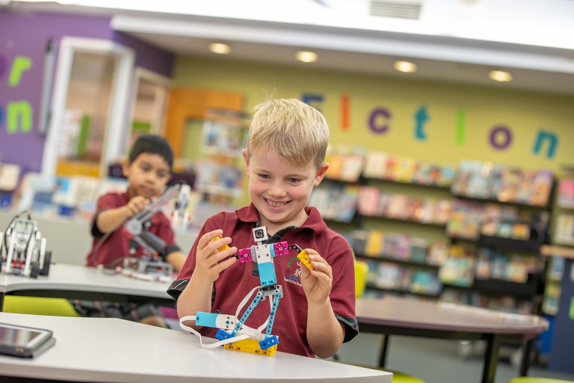 Primary school student assembling a small robotics model at a table, with a library in the background.
